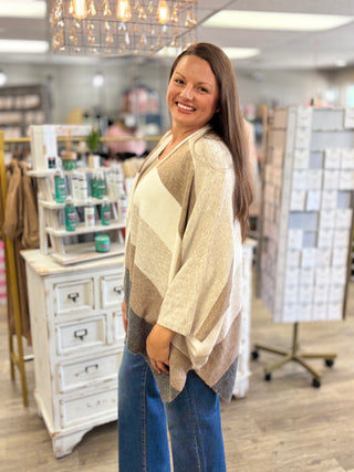 A woman modeling a beige and brown striped poncho cardigan in a boutique setting.