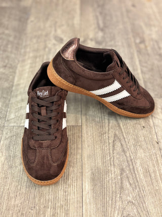 A pair of brown suede sneakers with white side stripes and gum soles, displayed on a light gray wood floor. One shoe is upright and the other is tilted on top of it.