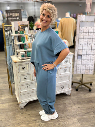 A woman with short blonde hair stands in a retail store wearing a matching dusty blue lounge set consisting of a short-sleeve top and jogger pants. She is posing in front of white wooden display cabinets.