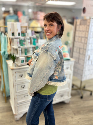 A woman posing in a boutique wearing a light-wash denim jacket with colorful floral embroidery on the back and white lace trim at the cuffs, layered over a green top and blue jeans.