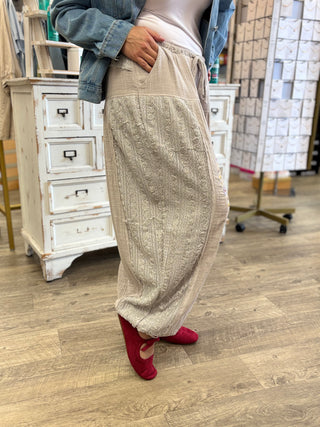 A person wearing loose, textured beige knit pants with a wide waistband, paired with a light top, denim jacket, and red slip-on shoes, standing in a retail store with wood floors and white cabinets.