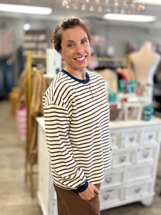 A smiling woman with short brown hair stands in a boutique wearing a cream and dark brown horizontally striped long-sleeve shirt with a navy blue crew neck and cuffs, paired with brown pants.