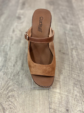A single tan suede slide sandal with an ankle strap and a low wedge heel, displayed on a clear acrylic block over a gray wood-look floor. 