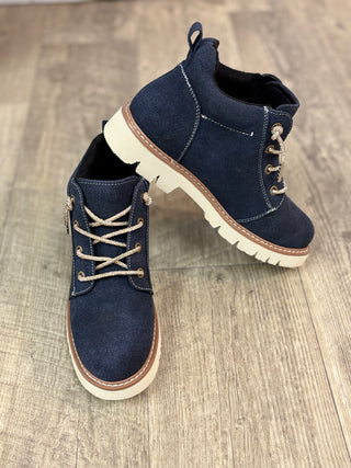 A pair of navy blue suede lace-up ankle boots with beige laces and thick off-white soles, displayed on a light brown wood floor.