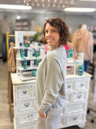 A woman with short brown curly hair smiling and looking over her shoulder in a boutique, wearing a light gray heathered sweatshirt and matching sweatpants. The background shows a white wooden cabinet with toiletries and mannequins in a softly blurred store setting.