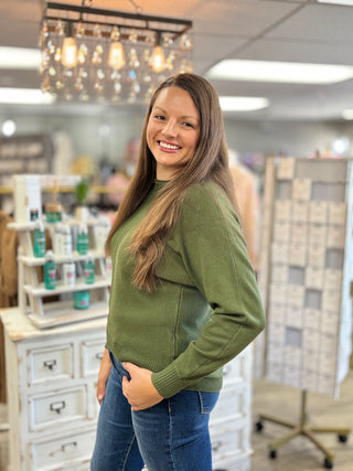 A woman with long brown hair smiling in a retail store, wearing an olive green knit sweater and blue jeans.