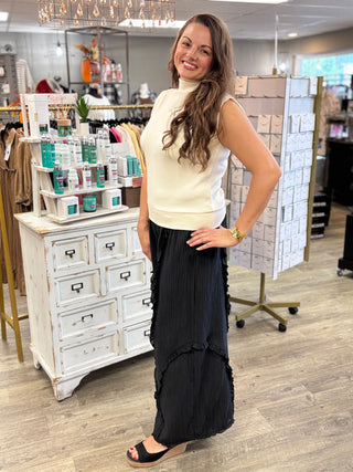 Model wearing a cream colored mock neck, sleeveless top with black textured pants