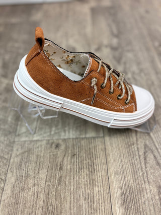 A brown suede low-top sneaker with white laces and a thick white sole, displayed on a clear acrylic stand over a gray wood-look floor.