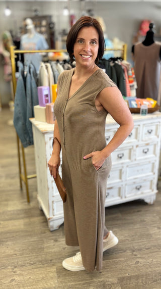 A woman modeling a sleeveless, ribbed knit jump suit in a taupe-brown color, standing inside a clothing boutique.