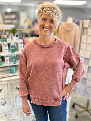 A smiling woman with short blonde hair wearing a dark rust long-sleeve crewneck sweatshirt and blue jeans, standing in a retail store setting.