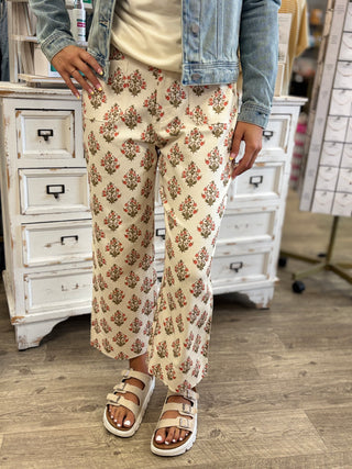 A person wearing cream wide-leg pants with a rust and green floral pattern, styled with a light blue denim jacket, a cream top, and beige slide sandals in a boutique setting.