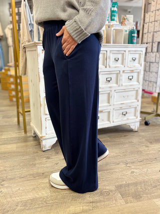 A person wearing navy wide-leg pants with a gray knit sweater and white sneakers, standing in a retail store with wooden floors and white furniture in the background.