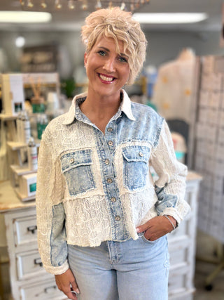 A woman with short blonde hair stands in a boutique wearing a light blue denim jacket with cream lace applique panels and light blue jeans.