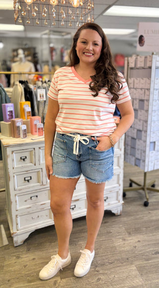 Model is wearing striped top with medium wash, fray hem shorts with tie cord detail. Shorts feature square front pockets.