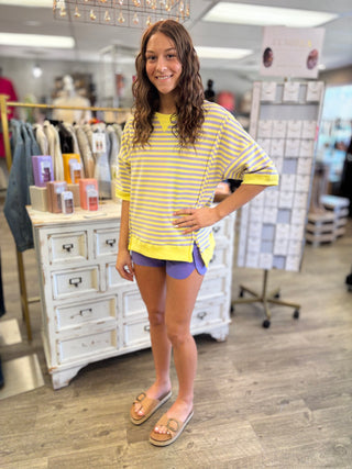 Model wearing a yellow and lavender striped shirt and purple shorts in a store setting.