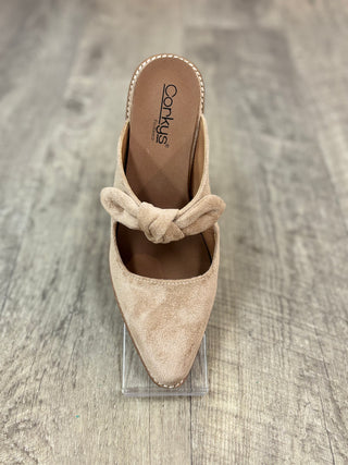 A beige suede pointed-toe mule slipper with a knotted bow detail across the instep, displayed on a clear acrylic stand over a gray wood-look floor.