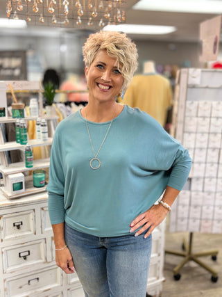 A smiling woman with short blonde hair stands in a boutique wearing a dusty teal blue long-sleeve top, blue jeans, and a silver necklace with a circular pendant.