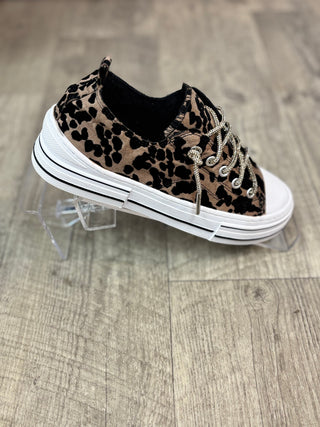 A low-top sneaker with a brown and black leopard print upper, silver metal eyelets, and a thick white platform sole, displayed on clear acrylic stands over a light wood floor.