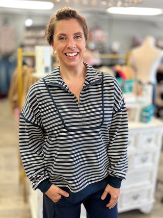 A smiling woman with short brown hair stands in a retail store wearing a navy and cream horizontal striped long-sleeve quarter-zip pullover with dark navy pants.