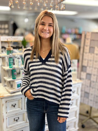 A smiling young woman with blonde hair stands in a boutique shop wearing a navy and cream wavy knit sweater and blue jeans.
