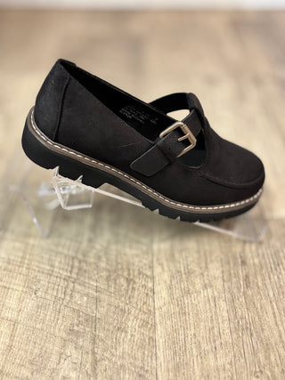A single black Mary Jane-style shoe with a silver buckle, displayed on a clear acrylic stand over a light wood floor.