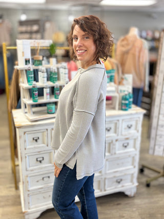 A woman with short brown hair smiling and looking over her shoulder, wearing a light gray ribbed long-sleeve top and blue jeans, standing in a retail store setting with white wooden display cabinets behind her.