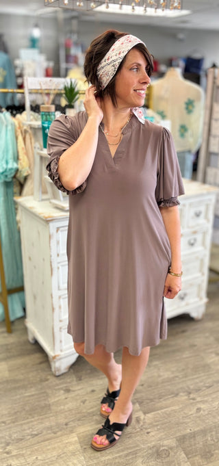 A woman modeling a short-sleeve, V-neck shift dress in a muted taupe-brown color inside a retail boutique. She is wearing a patterned headband and black slide sandals.