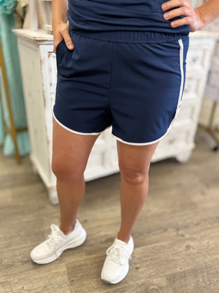 Model wearing navy shorts with white trim and white sneakers indoors.