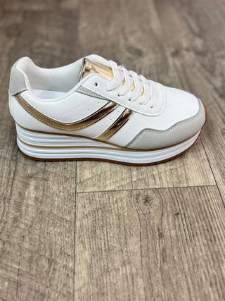 A white low-top platform sneaker with metallic gold accents on the side stripes and tongue, resting on a brown wood-look floor.