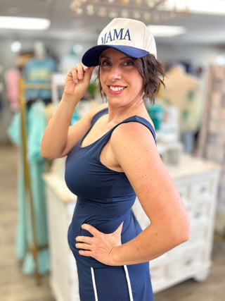 Woman wearing a navy tank top and blue pants with a 'MAMA' cap in a store setting.