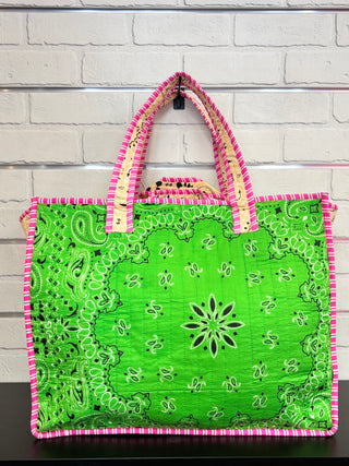 A green quilted tote bag with a paisley bandana pattern and pink and cream striped handles, hanging against a white brick wall.