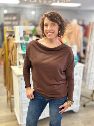A woman with short brown hair smiling in a clothing boutique, wearing a brown long-sleeve boat-neck top and blue jeans.