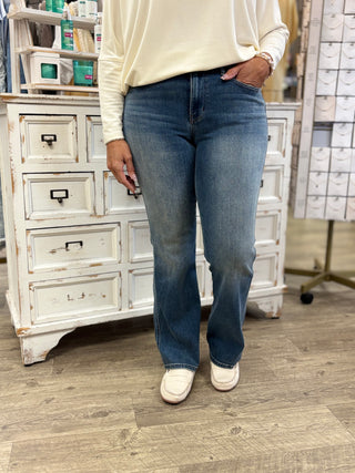 A person wearing medium-wash blue jeans with a slight flare, paired with a cream long-sleeve top and white slip-on shoes, standing in a store in front of a white wooden cabinet.
