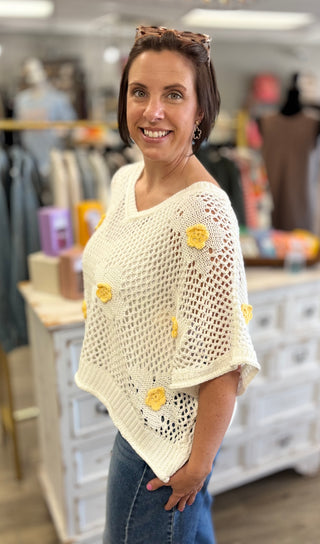 Woman wearing a light-colored crocheted poncho with yellow flowers in a store setting