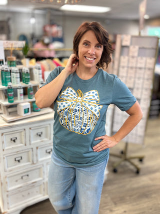 A woman posing in a retail store wearing a slate blue t-shirt with a large white, blue, and gold gift box graphic on the front, paired with light wash blue jeans.