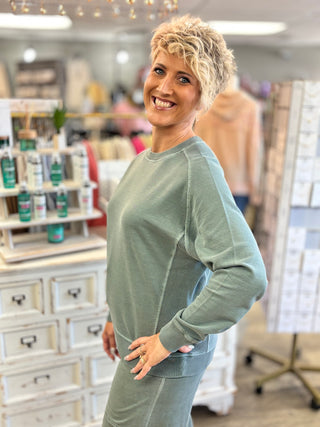 A smiling woman with short blonde hair models an ash jade long-sleeve crewneck sweatshirt and matching sweatpants inside a retail store.