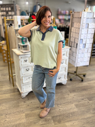 Model wearing an oversized top in a sage green color with blue trim and button details, paired with light wash denim and taupe Mary Jane style shoes