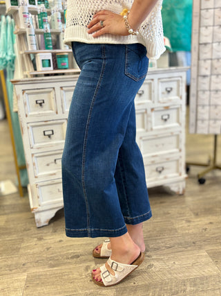 A person wearing a pair of dark blue cropped wide-leg denim jeans, paired with a cream knit top and beige slide sandals, standing in a boutique setting.