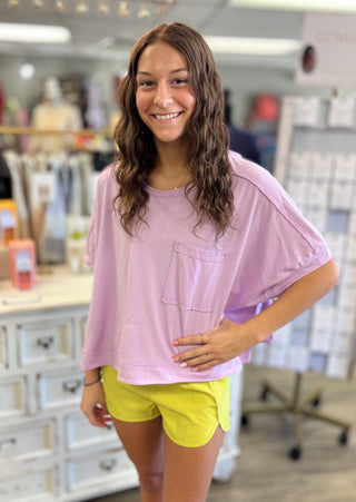 Model wearing a light purple oversized shirt and yellow shorts in an indoor setting.