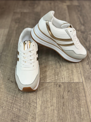 A pair of white low-top sneakers with metallic gold side stripes and a brown gum outsole, displayed on a rustic wood floor.