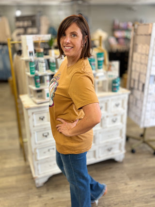 A woman with short brown hair stands in a retail store interior wearing a mustard yellow graphic t-shirt and blue jeans, looking back over her shoulder. White display cabinets and shelves with various products are visible in the background.