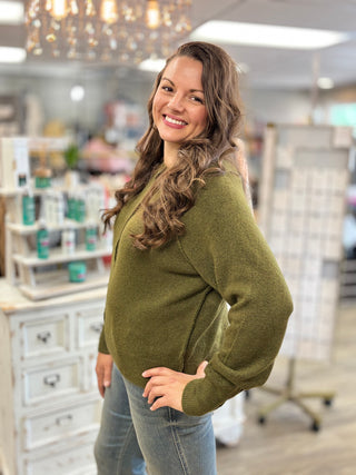 A woman with long wavy brown hair smiling and posing in a cozy olive green knit sweater and blue jeans inside a boutique store.