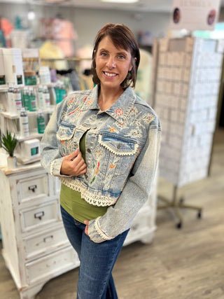 A woman posing in a boutique wearing a light-wash blue denim jacket with colorful floral embroidery and lace trim over a green top and blue jeans.
