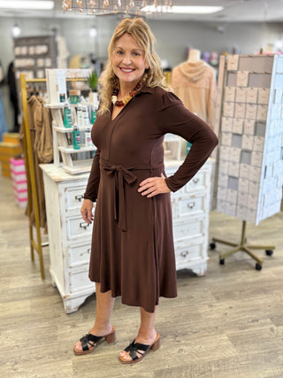 A woman modeling a long-sleeve brown midi dress with a tie waist, standing in a boutique store with white furniture and clothing racks in the background.