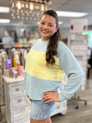 A woman modeling a color-blocked long-sleeve knit top with a light blue base, a wide pale yellow chest panel, and a matching light blue pocket, paired with denim shorts in a retail store setting.