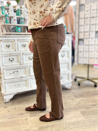 A person wearing brown straight-leg jeans, a cream floral knit sweater, and brown Mary Jane flats, standing in a boutique setting with a white wooden cabinet in the background.