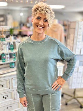 A smiling woman with short blonde hair stands in a retail store wearing a matching ash jade sweatshirt and sweatpants set.