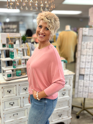 A smiling woman with short blonde hair stands in a boutique store wearing a pink long-sleeve top and blue jeans, with shelves of products and mannequins in the background.