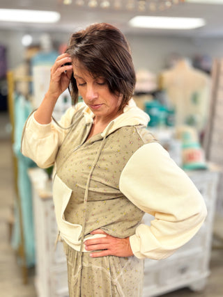 A woman wearing a beige and cream hooded sweatshirt dress with a small floral pattern, standing in a clothing boutique.