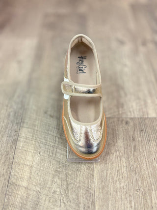 A single metallic gold Mary Jane shoe with a hook-and-loop strap, displayed on a clear stand over a light wood floor.